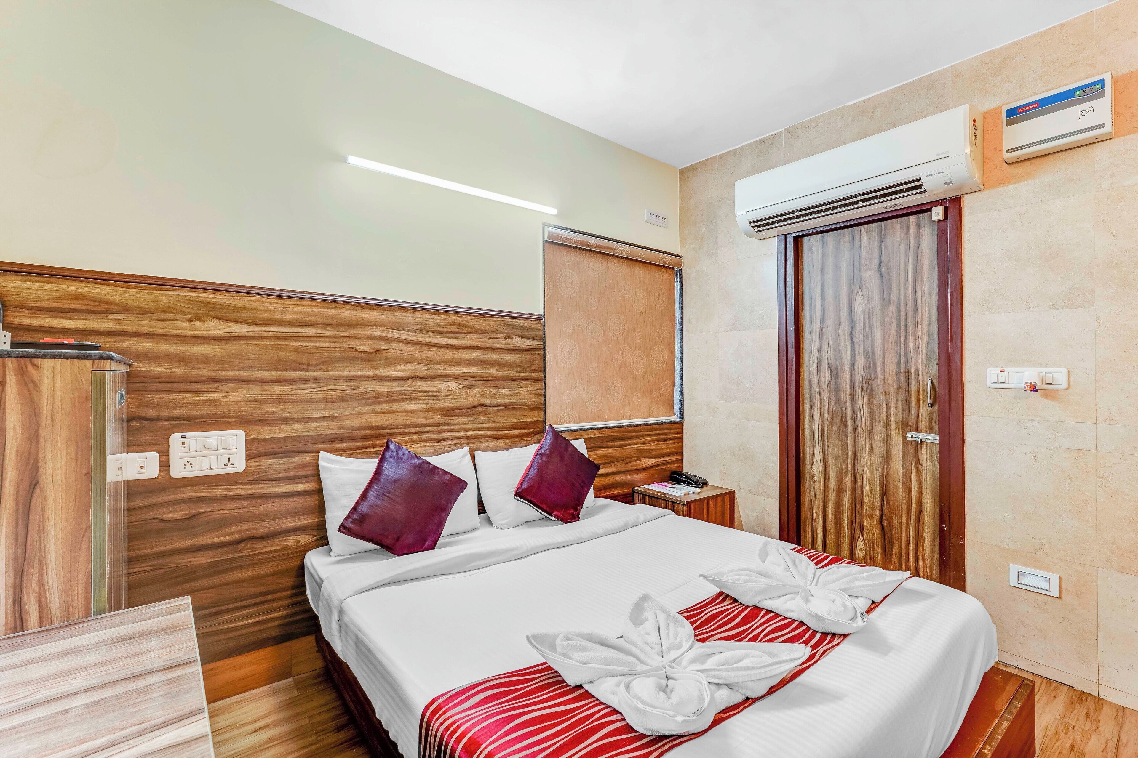 A hotel room with a wooden headboard, a bed with white bedding and red accents, a nightstand with a phone and a lamp, and an air conditioning unit on the wall.
