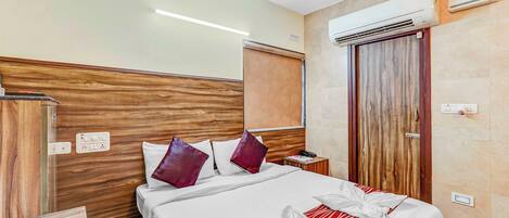 A hotel room with a wooden headboard, a bed with white bedding and red accents, a nightstand with a phone and a lamp, and an air conditioning unit on the wall.