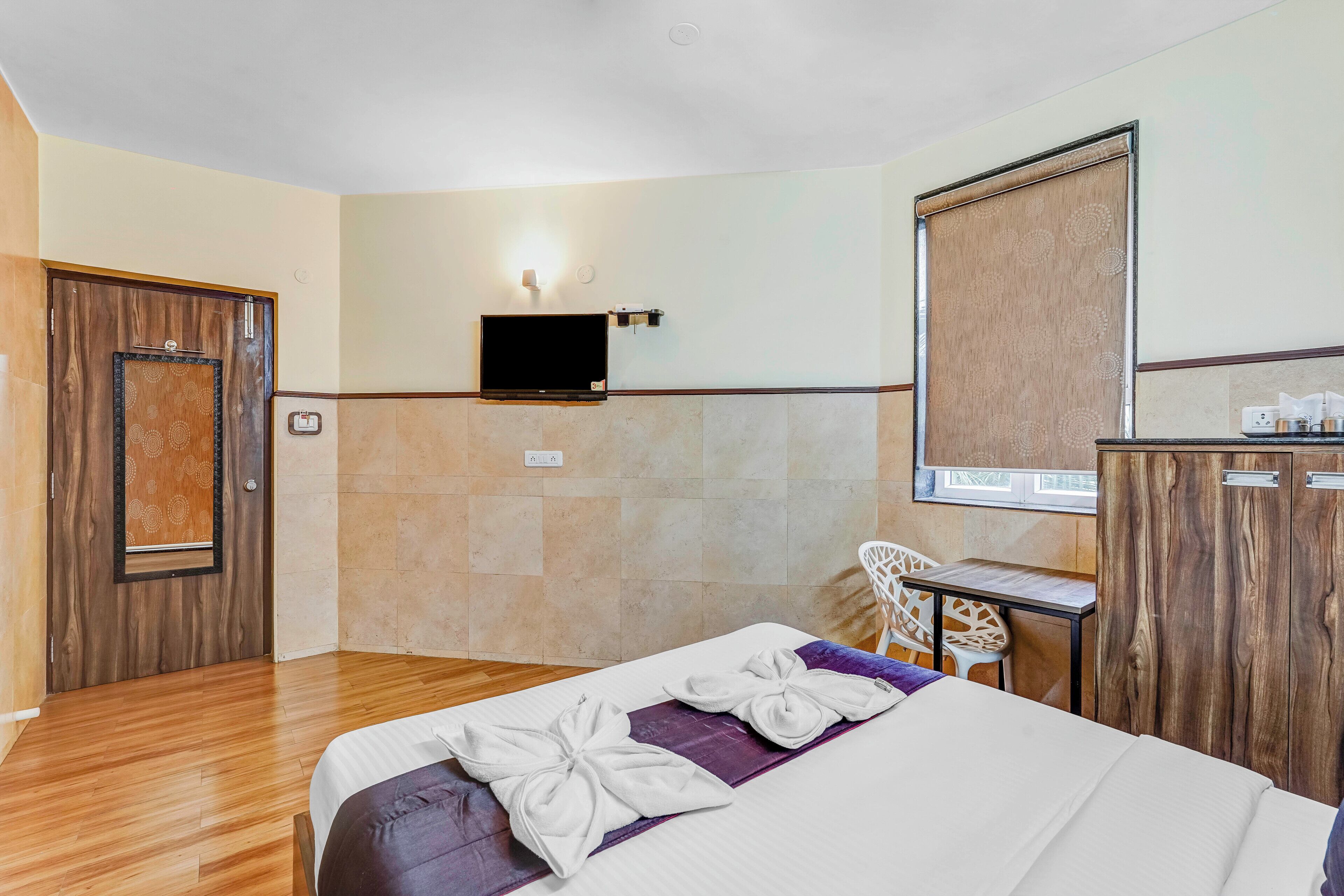 A hotel room with a bed, a TV mounted on the wall, a desk with a chair, and a wardrobe.