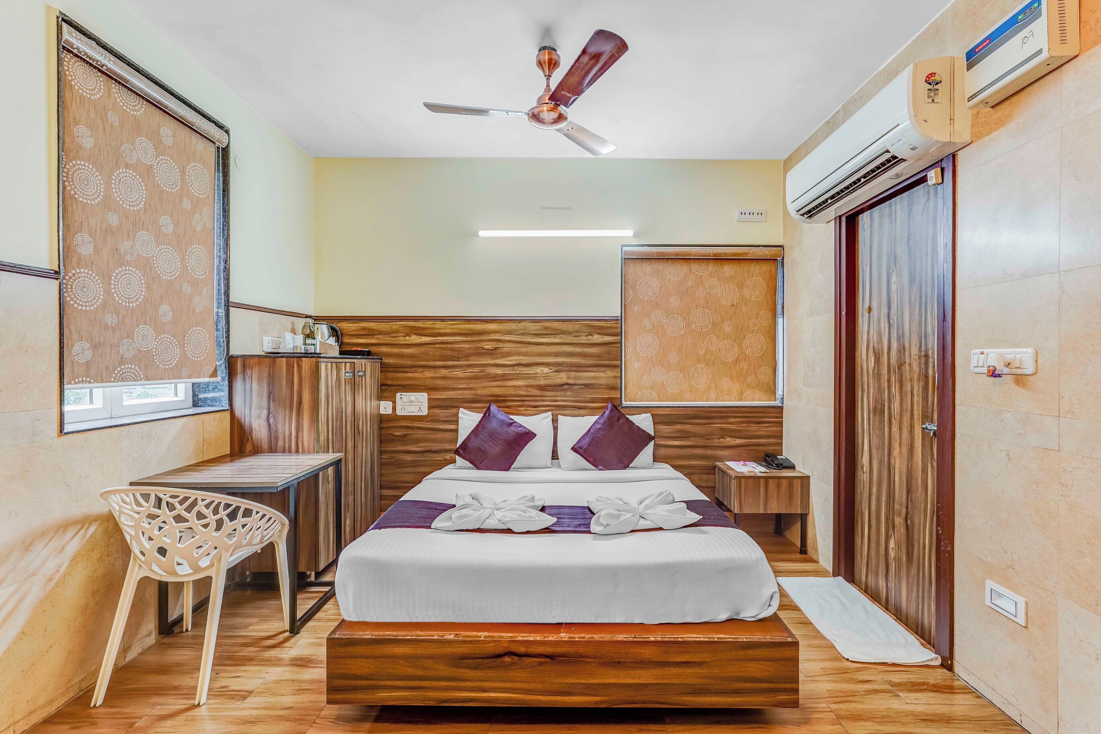 Une chambre d’hôtel avec un lit en bois, un ventilateur de plafond, un appareil de climatisation, une table de chevet, une chaise et une armoire.