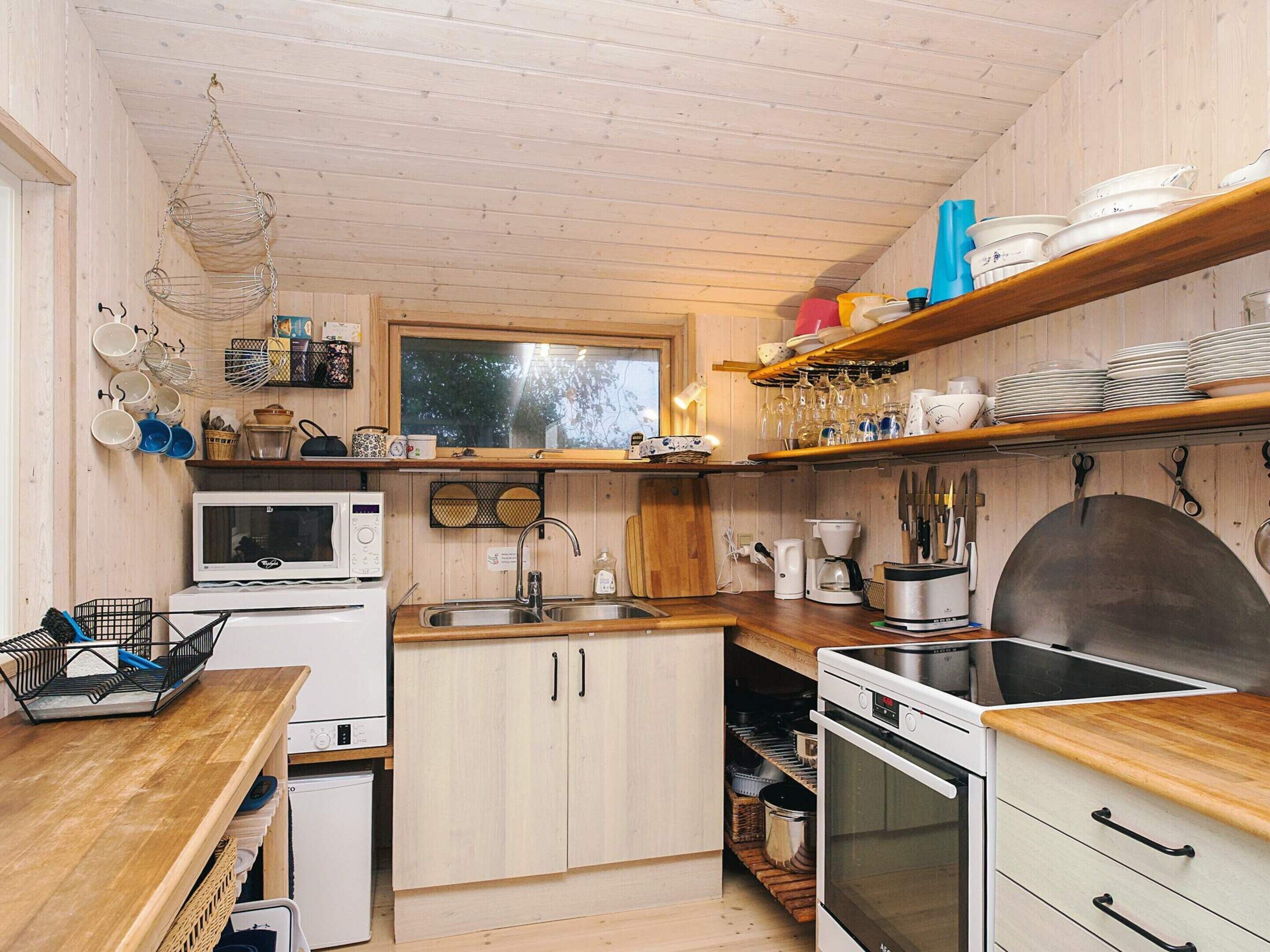 A compact kitchen with wooden cabinets, a microwave, a sink, a stove, and shelves with dishes and glasses.