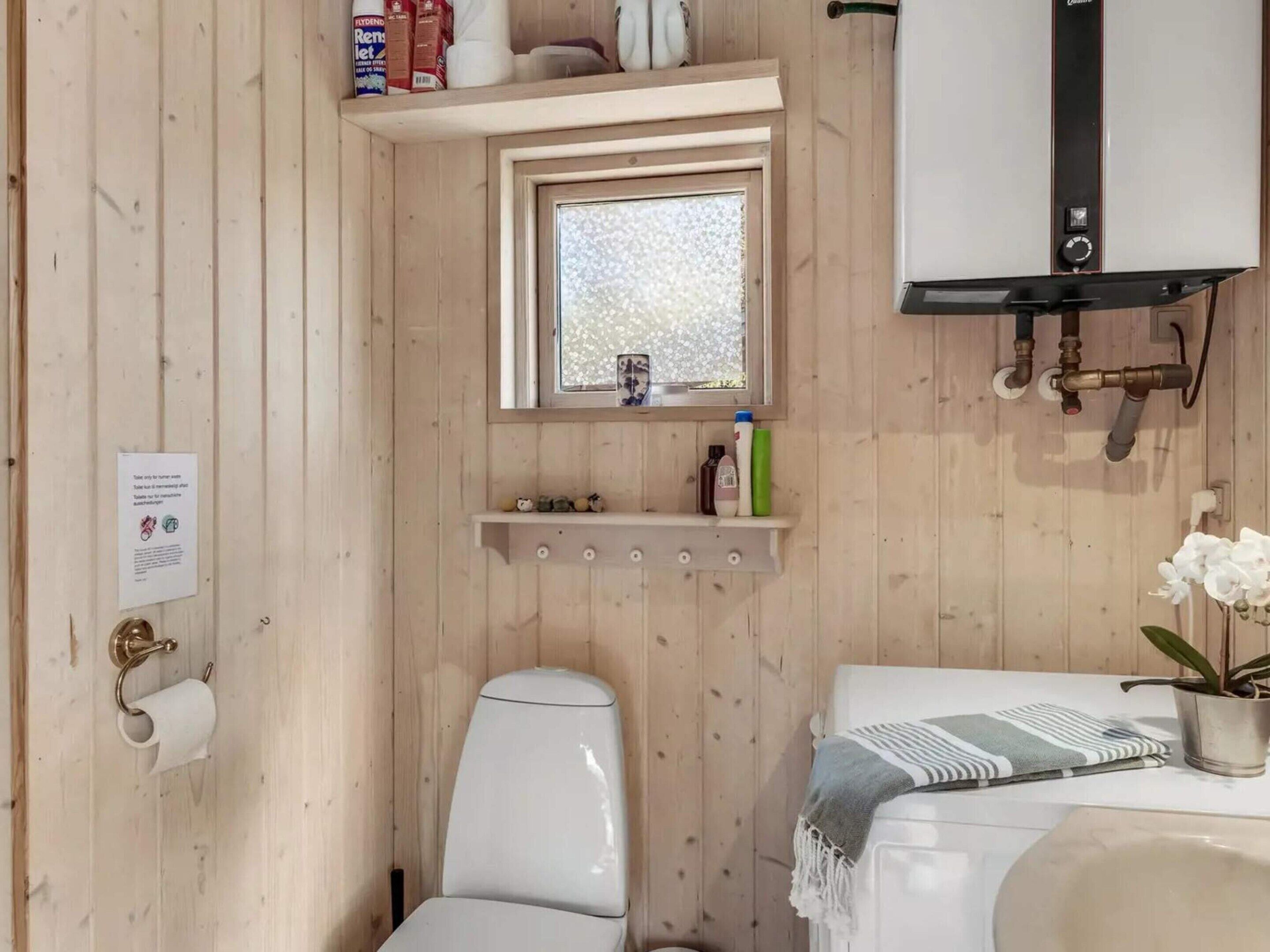 Et kompakt badeværelse med toilet, vask og vindue. Der er hylder over vinduet med toiletartikler og en lille plante.