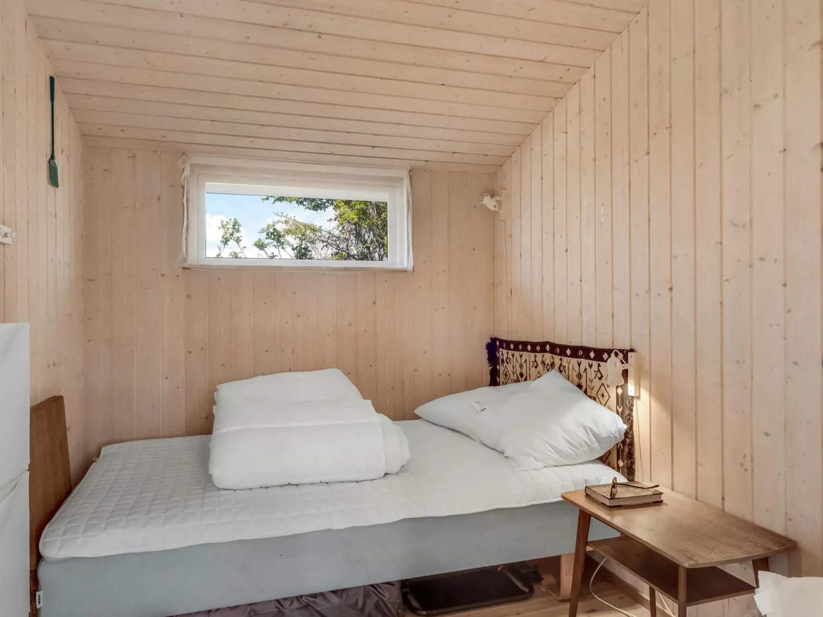 Pequeña habitación individual con cama, techo y paredes de madera, ventana con vista a árboles, mesita de noche con un libro y toma de corriente en la pared.