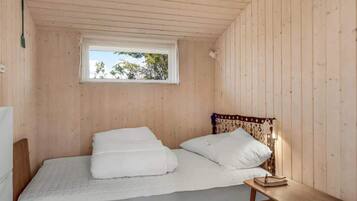 Pequeña habitación individual con cama, techo y paredes de madera, ventana con vista a árboles, mesita de noche con un libro y toma de corriente en la pared.