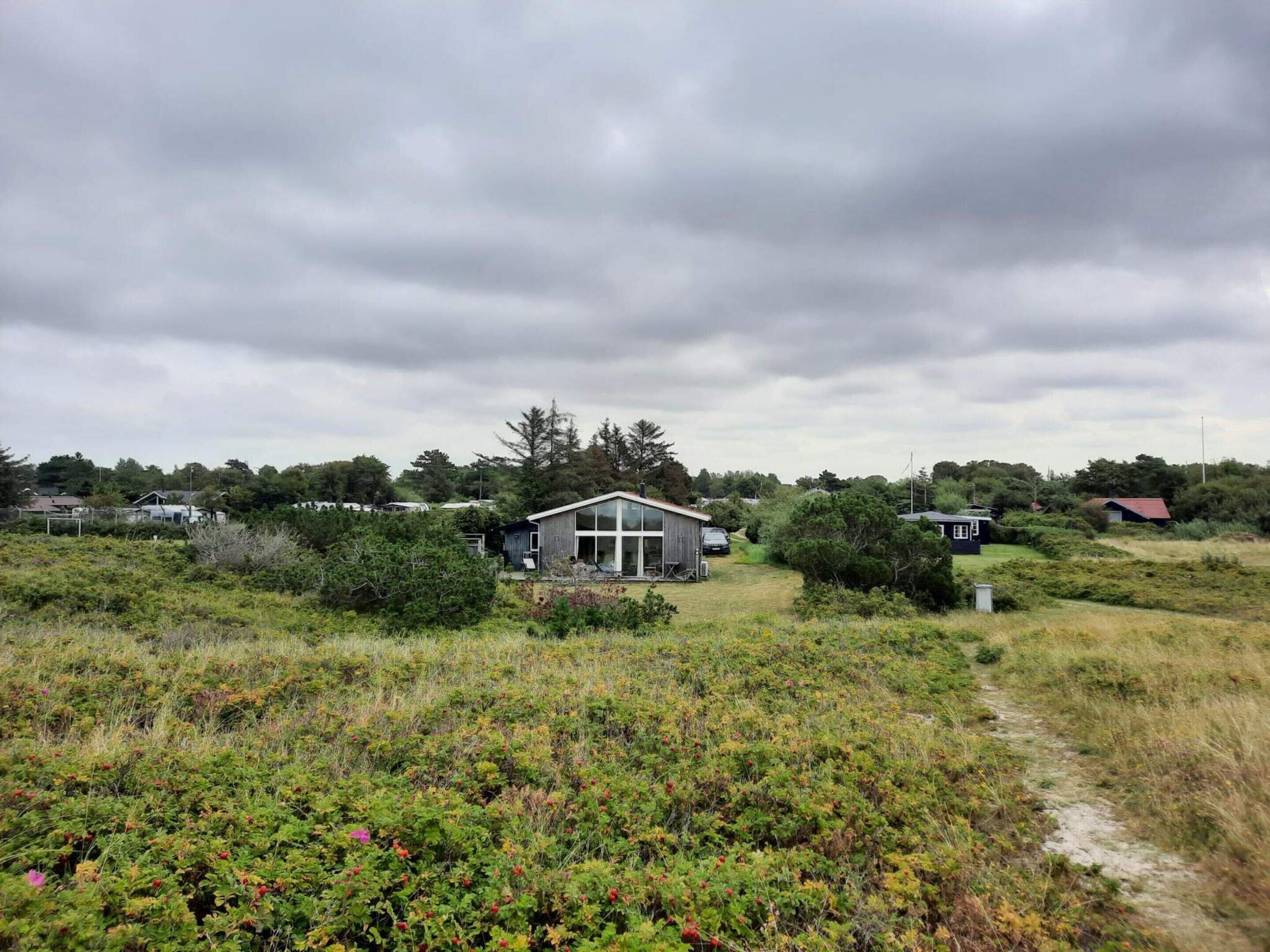 Une maison moderne dotée d’une grande façade vitrée, entourée d’un paysage naturel et accessible par un sentier.