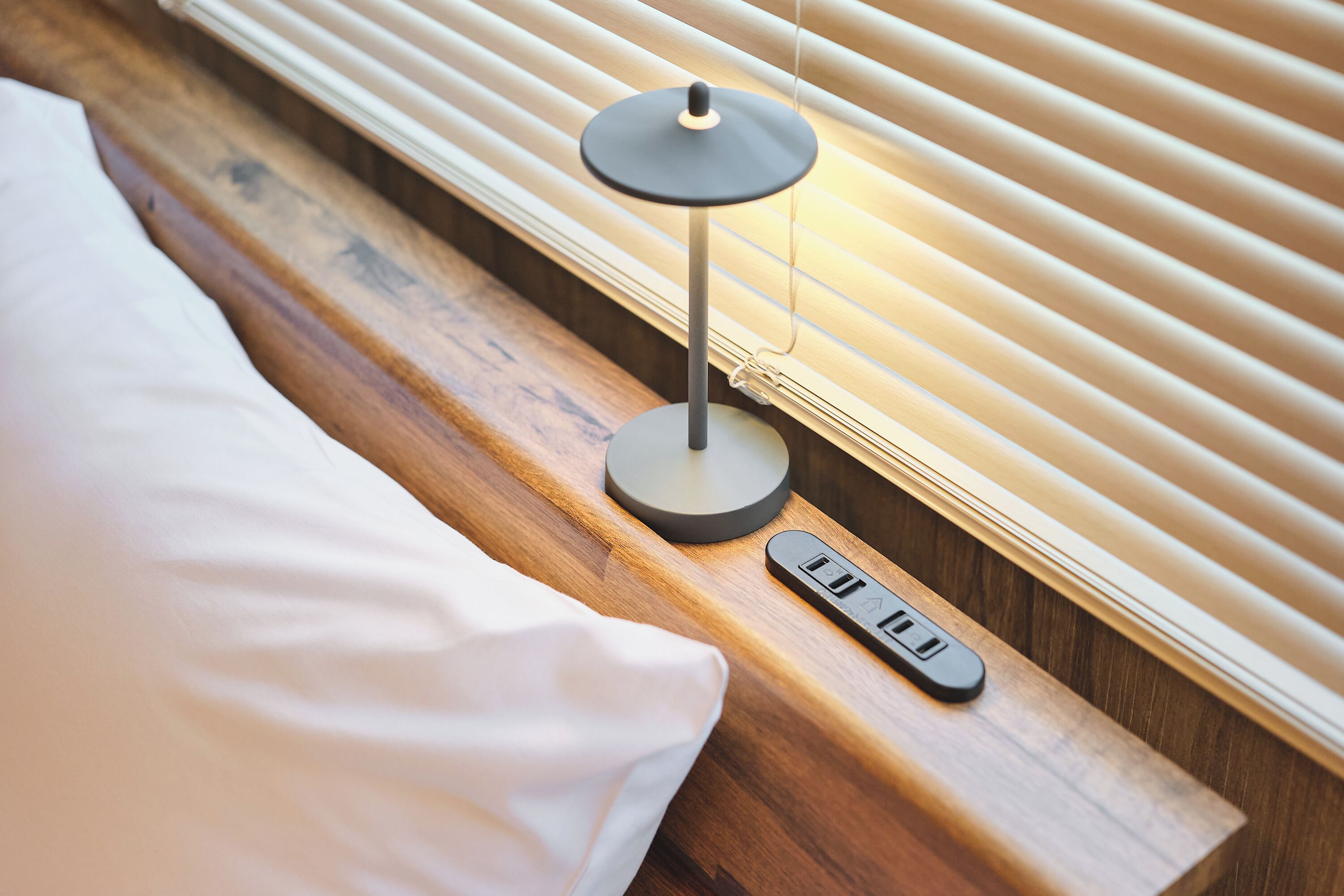 A bedside table with a lamp, a remote control, and a wooden headboard.