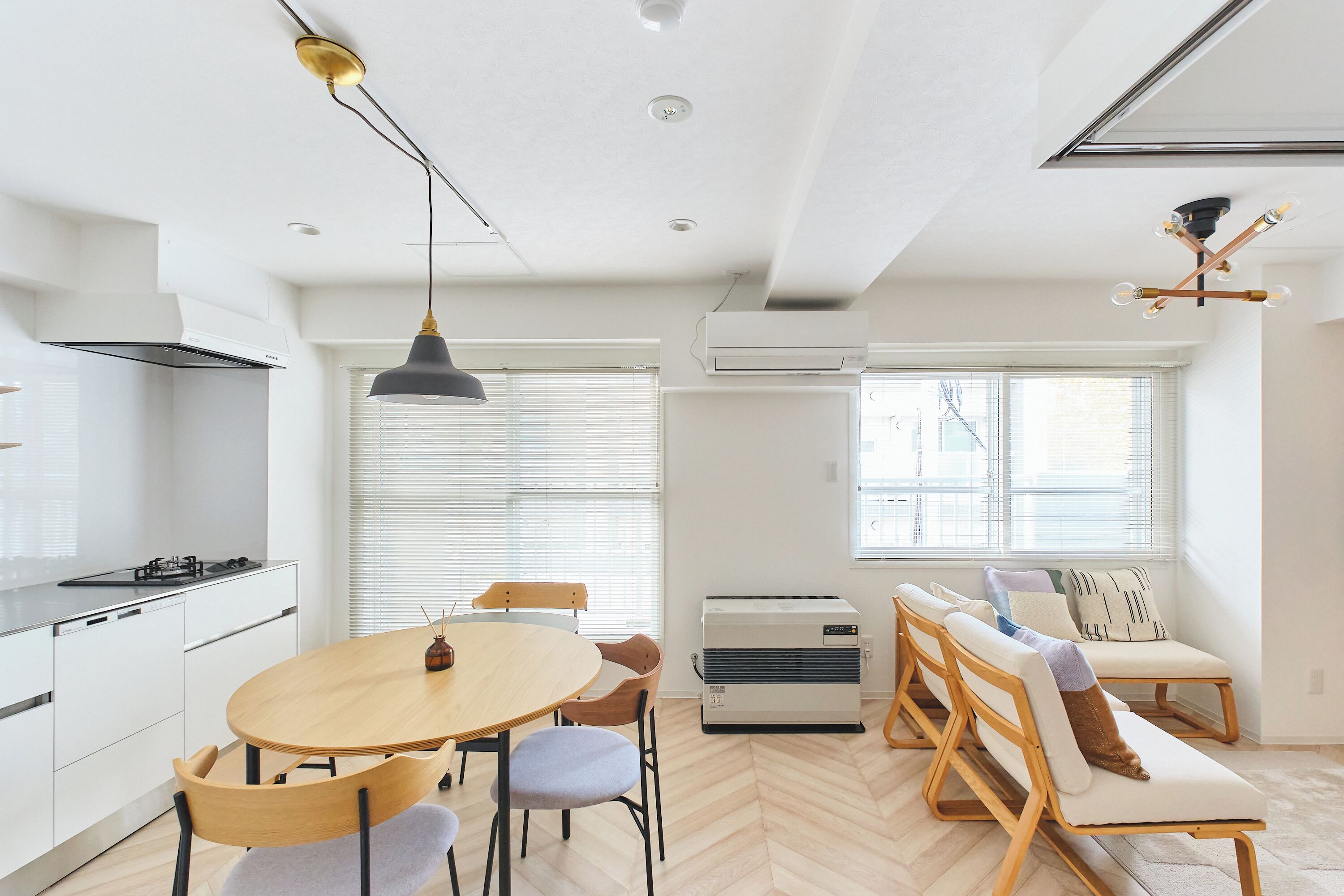 Eine moderne Küche mit weißen Schränken, einem runden Holztisch und einem eingebauten Herd. Es gibt eine Sitzecke mit einer Couch und Sesseln, sowie ein großes Fenster mit Jalousien.