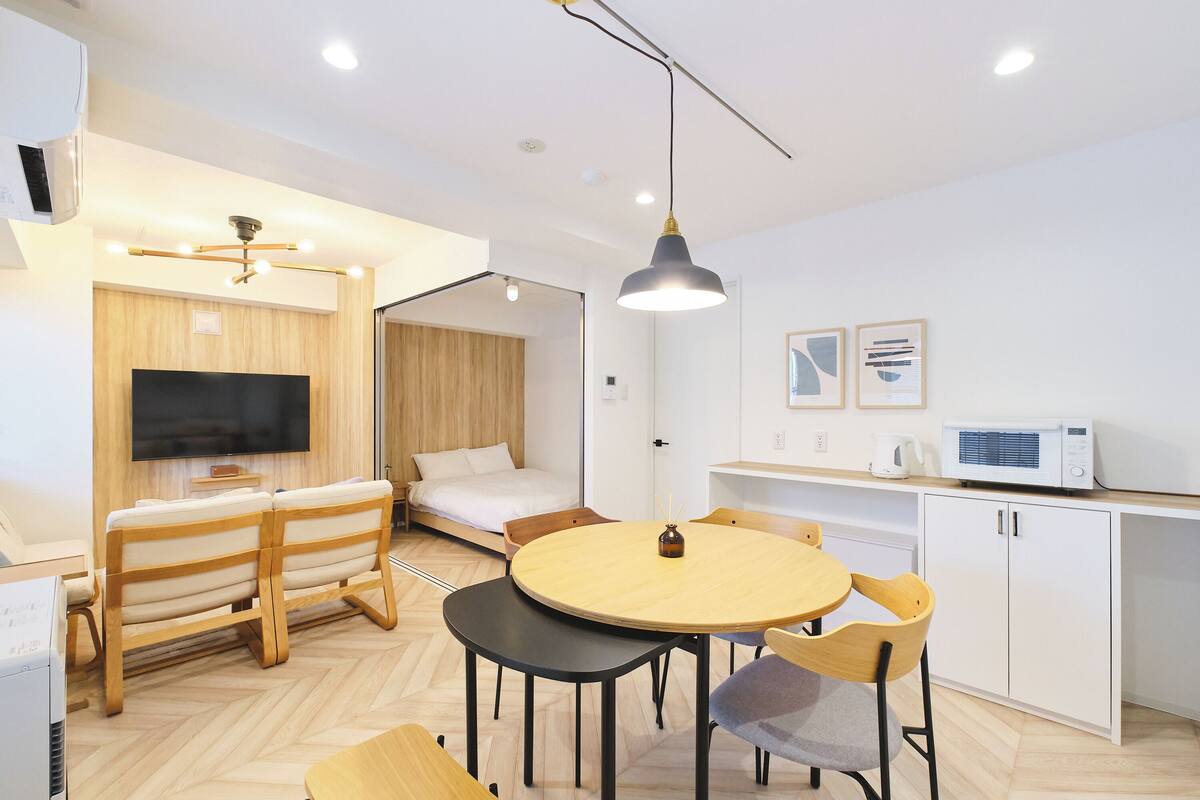 A modern, well-lit room with a round wooden table, two chairs, a sofa, a TV, and a microwave.