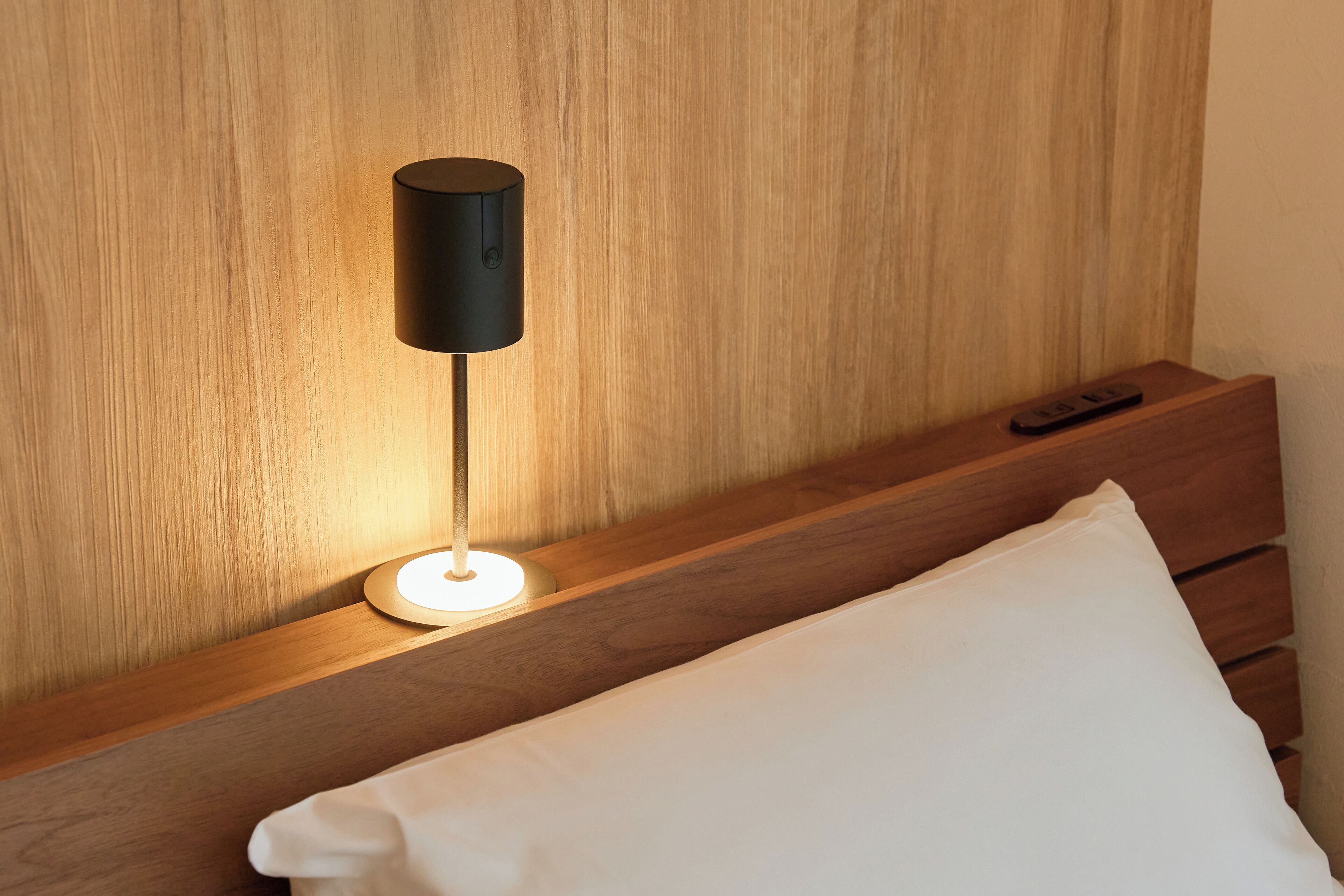A modern bedroom with a wooden headboard, a bedside table with a remote control, and a white pillow.