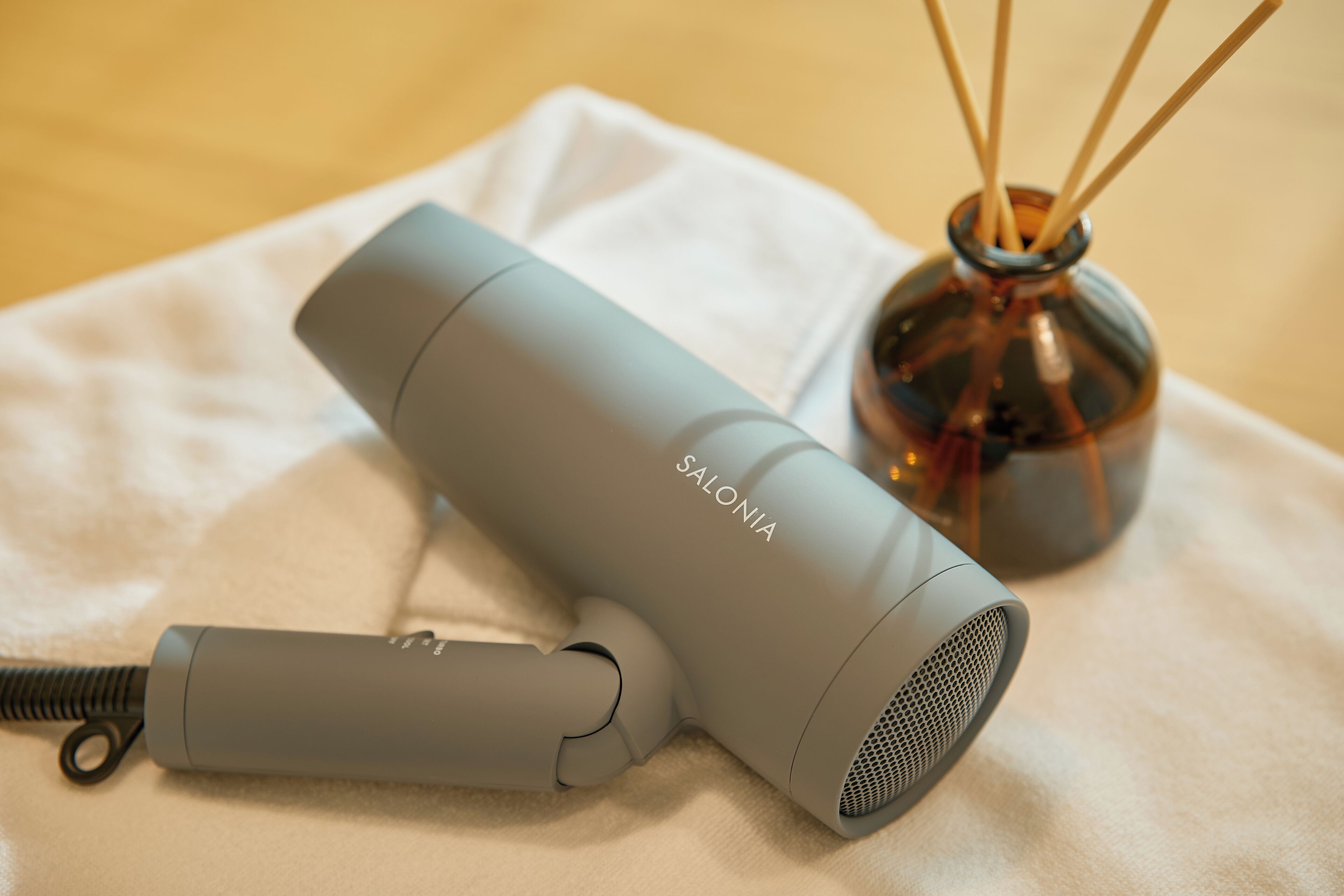 A gray hair dryer with a cord and a small bottle of reed diffuser with sticks on a white cloth.