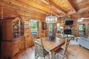 A wooden cabin interior with a dining table, chairs, a wooden cabinet, a stone fireplace, and a television. - Spacious 2BD Riverfront Cabin| Hot Tub| Steam shower| Fast WiFi| Private Retreat (Ellijay)
