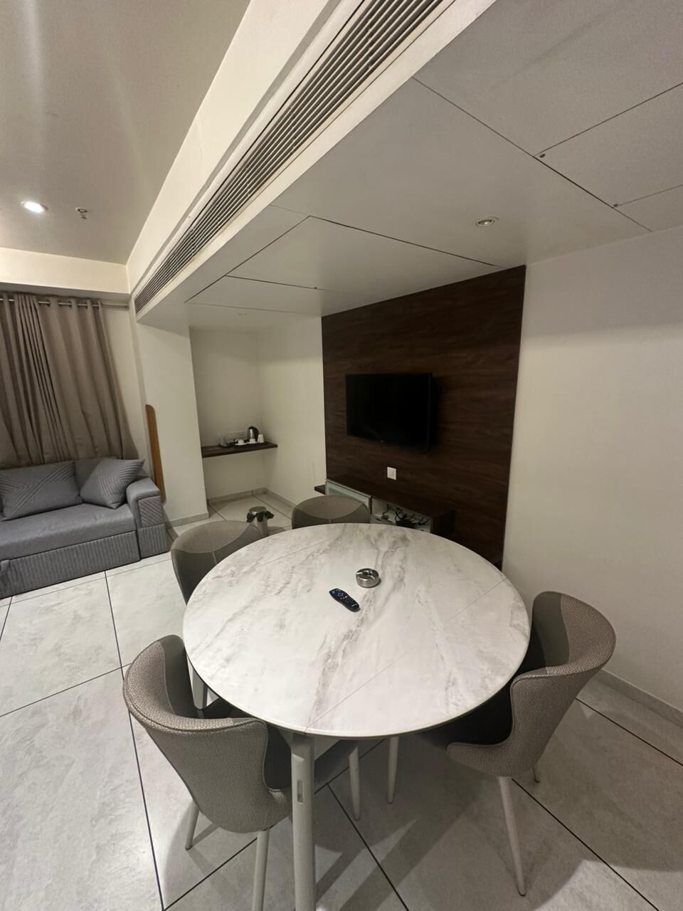 A modern living room with a round marble table, grey upholstered chairs, a grey sofa, a flat-screen TV mounted on a wooden wall, and a small shelf with items.