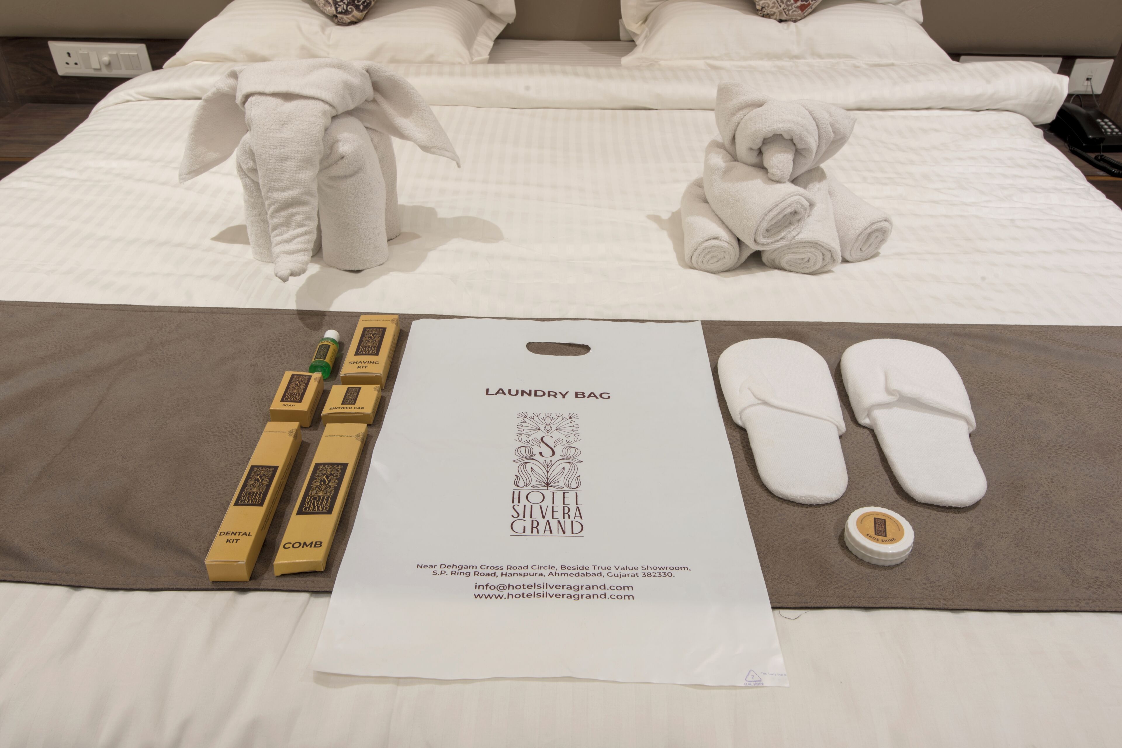 A neatly made bed with a white comforter, rolled towels, a dental kit, a comb, and a laundry bag on a brown bed runner.