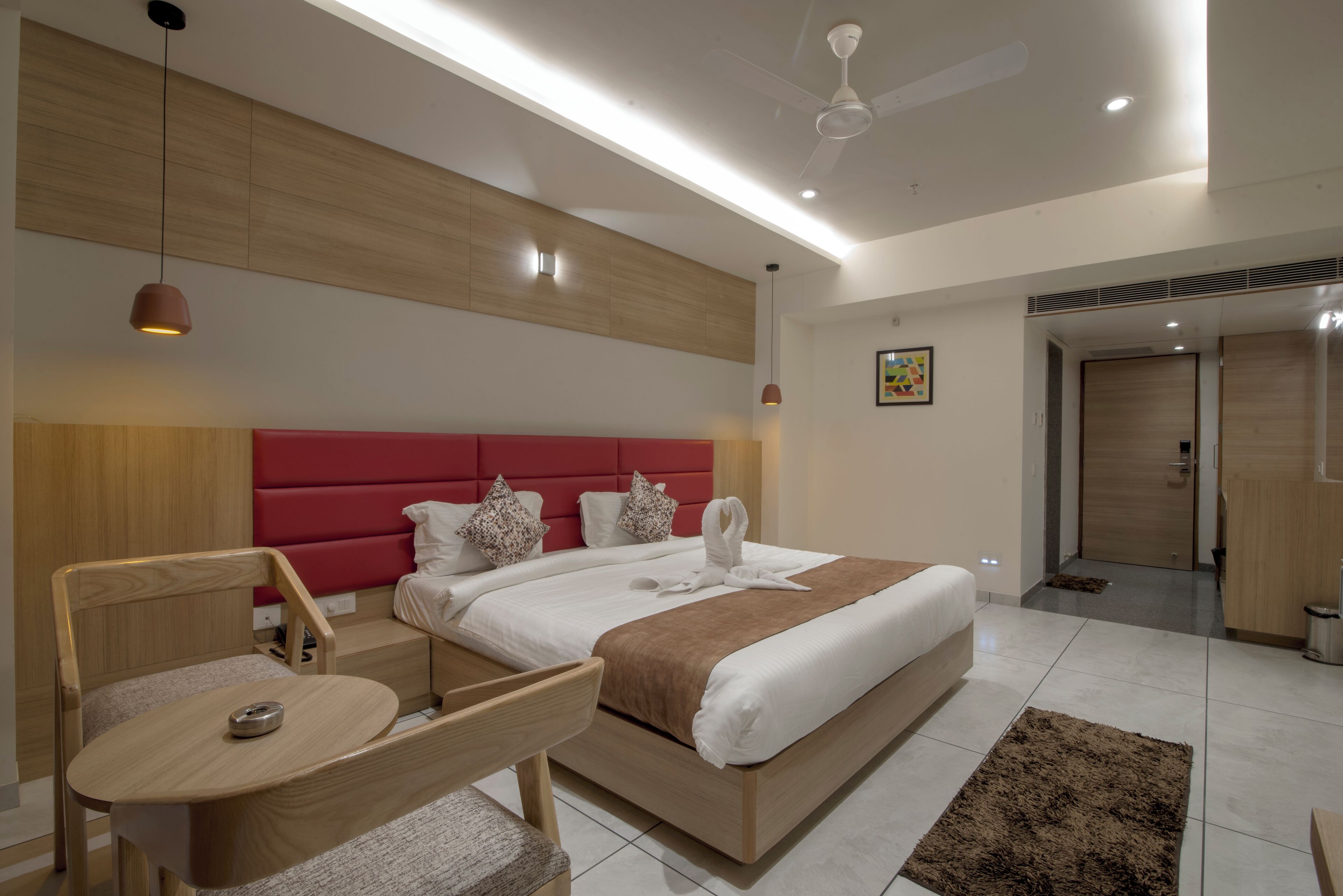 Ein modernes Hotelzimmer mit einem Holzkopfteil, einem Bett mit weißer und brauner Bettwäsche, einem kleinen Holztisch mit Stuhl und einem Deckenventilator.