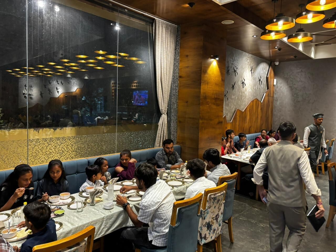 Restaurante com mesas postas para refeição, um garçom atendendo aos clientes e uma grande janela com vista para a paisagem urbana.