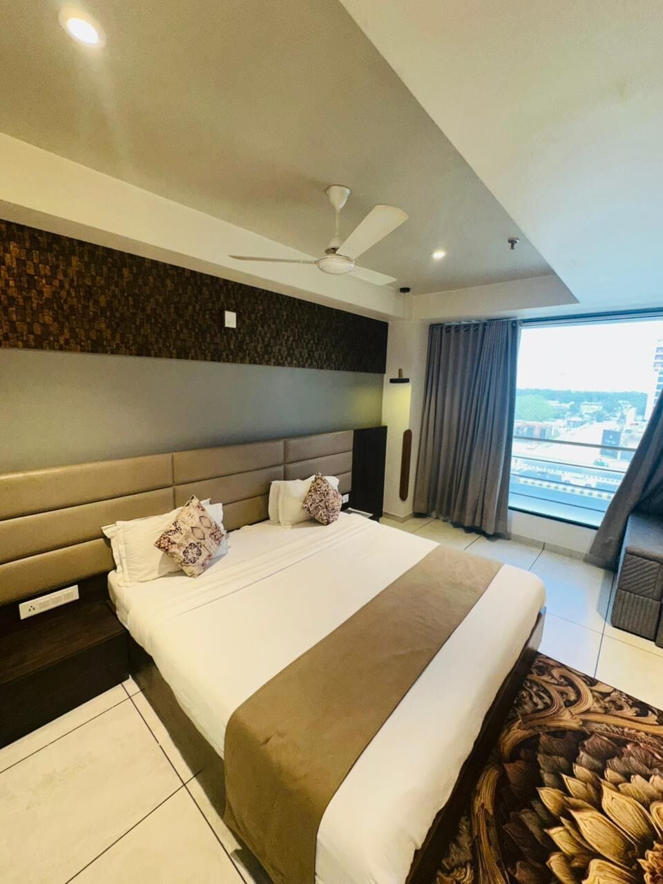A hotel room with a large bed, a ceiling fan, a view of the city, and a balcony.