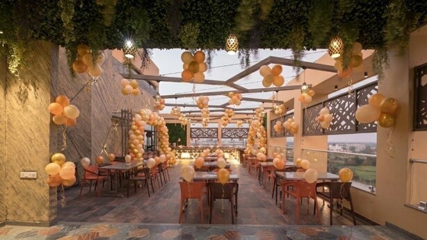 An indoor dining area with tables and chairs, a view through large windows, and decorative lighting. - Hotel Silvera Grand (Ahmedabad)