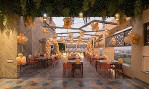 Een eetruimte binnenshuis met tafels en stoelen, een uitzicht door grote ramen en decoratieve verlichting.