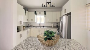 Una cocina moderna con armarios blancos, encimera de granito, refrigerador de acero inoxidable y una lámpara colgante.