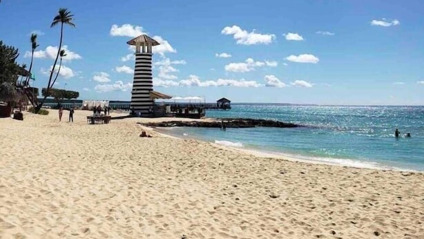 Una playa con un alto faro a rayas, un muelle que se adentra en el agua y palmeras.