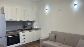 A modern kitchen with white cabinets, a built-in oven, and a grey countertop. There is a grey sofa and a small table with a chair in the living area. - SIKAN home By the sea (Butera)