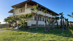 A two-story building with a flat roof, multiple balconies, and palm trees in the foreground. - SIKAN home By the sea (Butera)