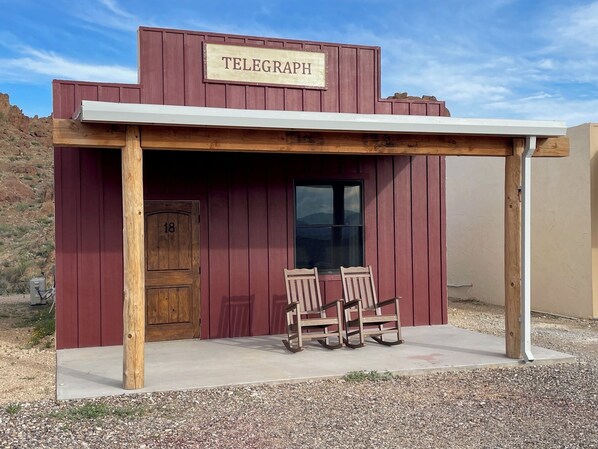 A rustic building labeled "TELEGRAPH" with a wooden door marked "18", two wooden rocking chairs, and a small window. - Ten Bits Ranch - Room # 18 - Telegraph (Terlingua)
