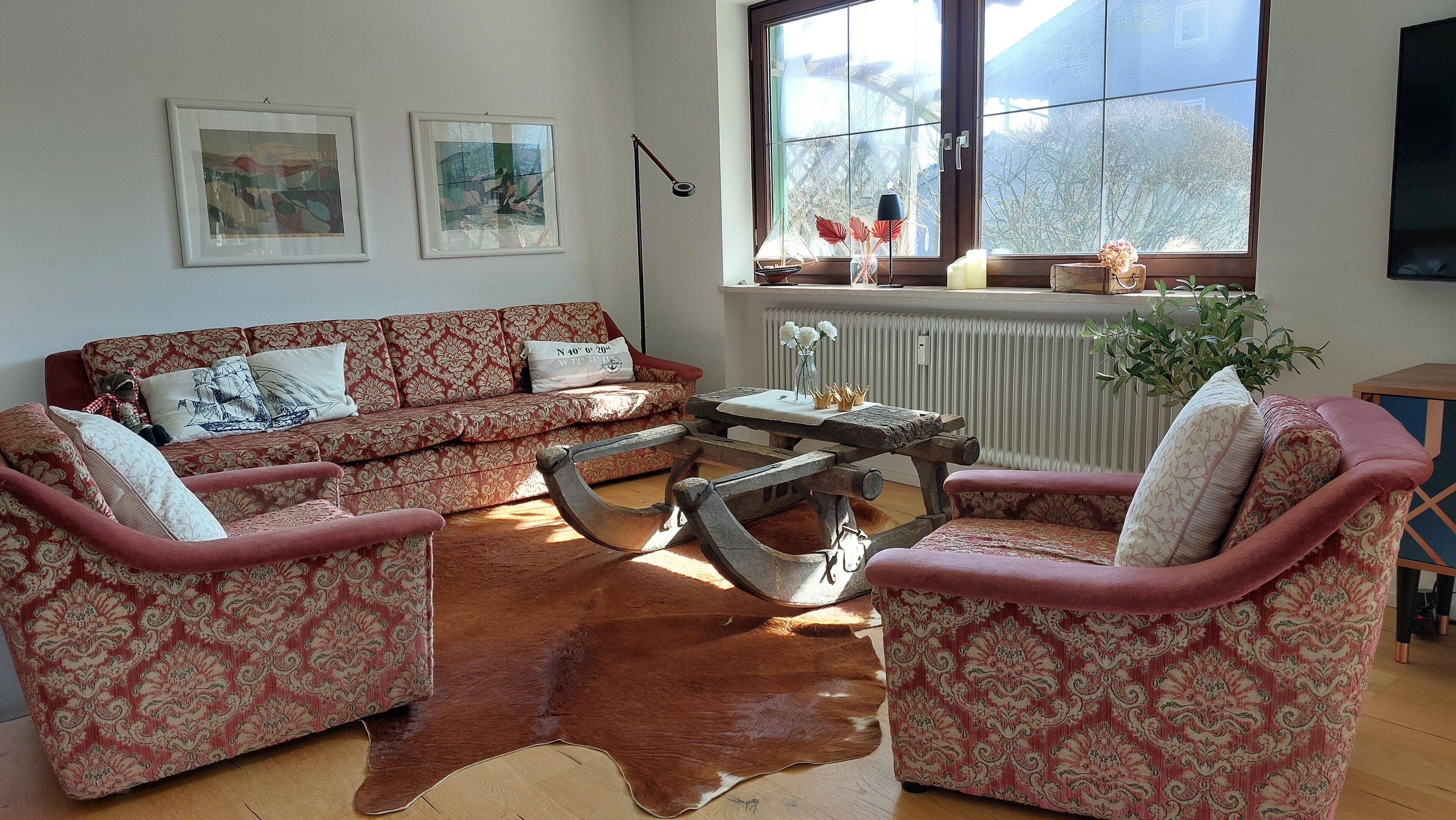 Ein Wohnzimmer mit einem gemusterten Sofa, zwei Sesseln, einem Couchtisch und einem großen Fenster mit Blick auf Bäume.