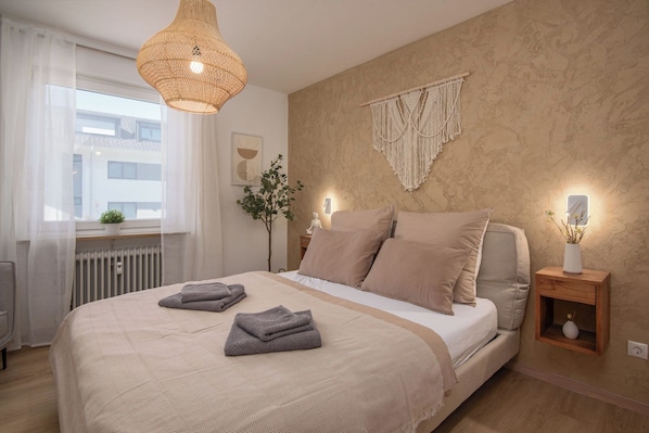A bedroom with a bed, bedside tables, a hanging light fixture, a macrame wall hanging, and a window with curtains.