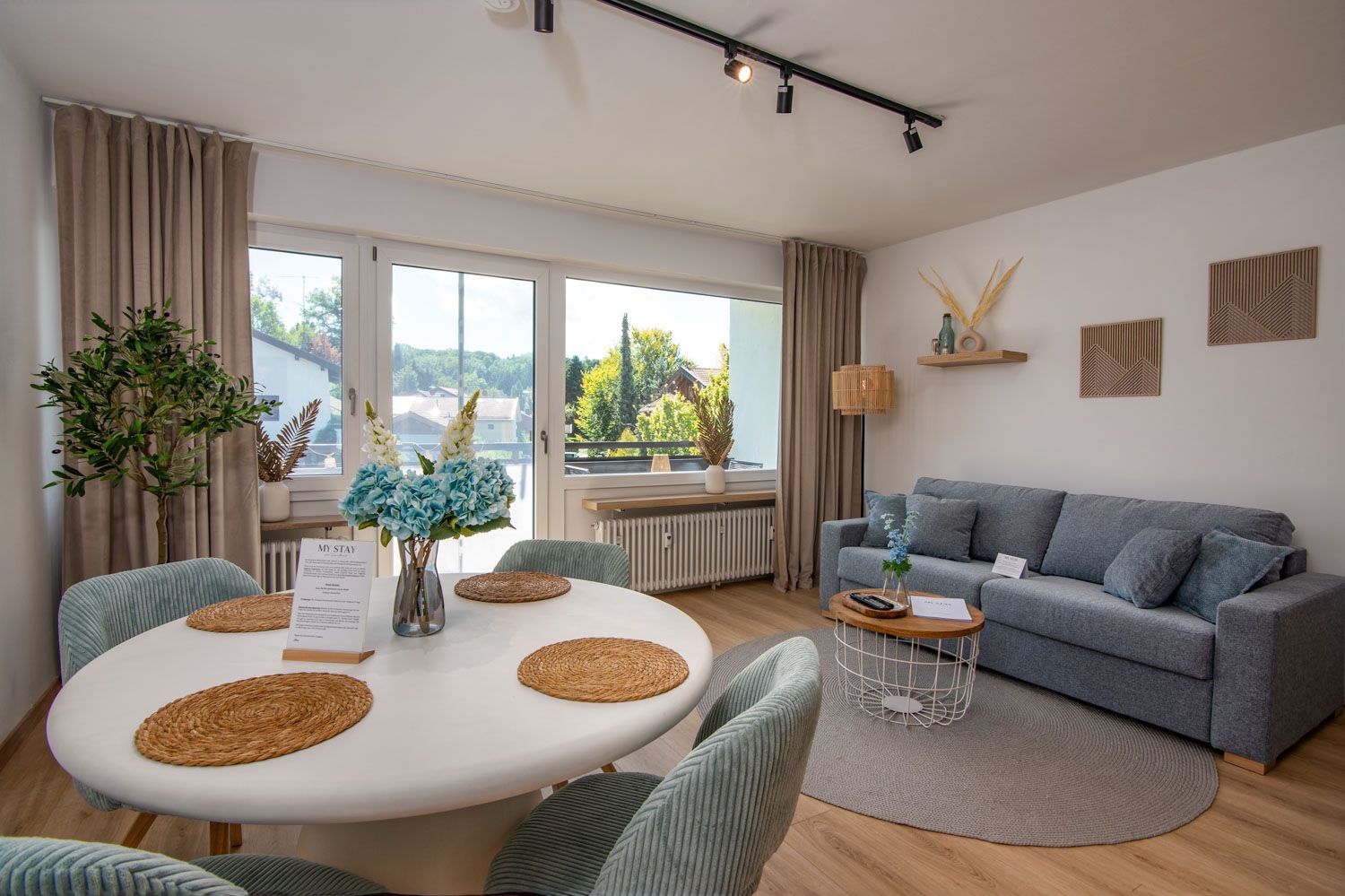 Un salon moderne avec un canapé gris, une table à manger ronde agrémentée de sets de table, et une vue sur la verdure à l’extérieur.