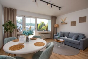 Een moderne woonkamer met een grijze bank, een ronde eettafel met plaatsmatten en uitzicht op groen buiten.