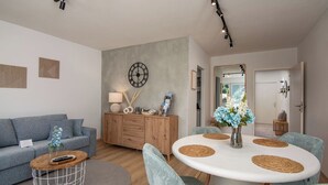A modern living room with a grey sofa, a wooden cabinet, a round dining table with woven placemats, and a large wall clock. - MY STAY - Relaxation on Lake Chiemsee in a central location in Prien (Prien am Chiemsee)