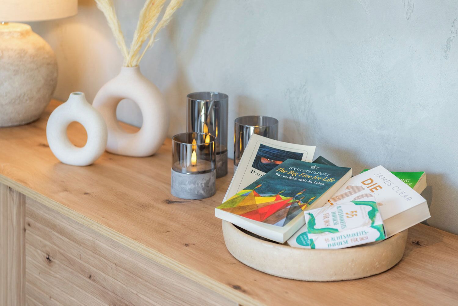 Ein Holztisch mit einer runden Holzschale, die Bücher, eine weiße Vase mit getrockneten Schilfhalmen und zwei zylindrische Glaswindlichter enthält.
