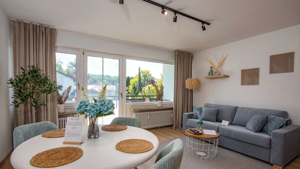 A modern living room with a grey sofa, a round dining table with placemats, and a view of greenery outside. - MY STAY - Relaxation on Lake Chiemsee in a central location in Prien (Prien am Chiemsee)