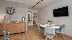 A modern living room with a wooden cabinet, a round table with four chairs, a wall clock, and a flat-screen TV mounted on the wall. - MY STAY - Relaxation on Lake Chiemsee in a central location in Prien (Prien am Chiemsee)