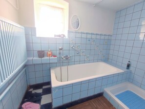 Baño con bañera, ducha y lavamanos. Hay una ventana con cortina y un estante con artículos de tocador.