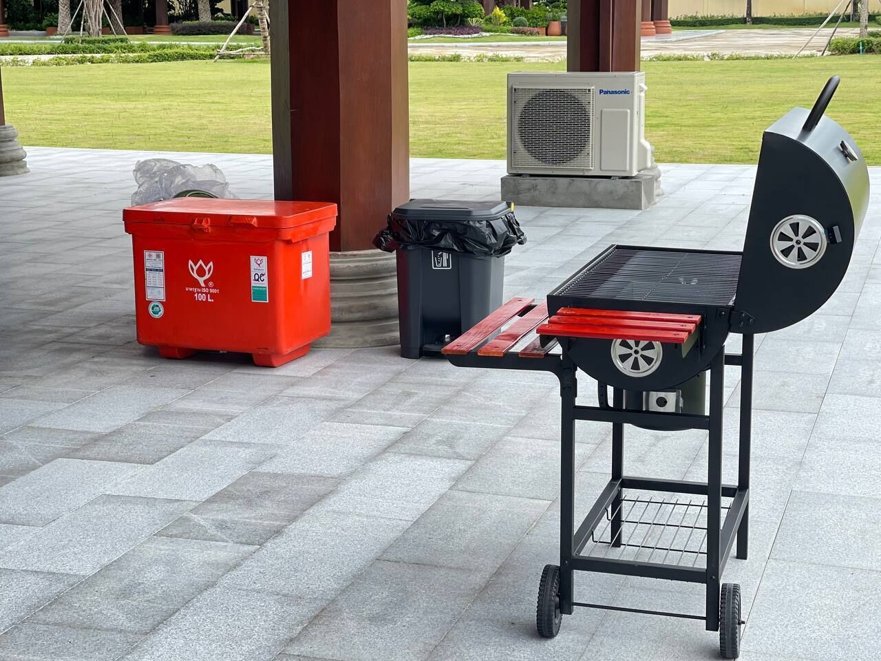 Un barbecue portable avec un plateau rouge et des roulettes, une poubelle rouge et une poubelle noire, le tout placé sur une surface extérieure pavée.
