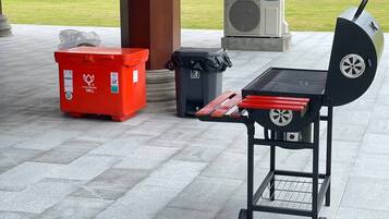 Un barbecue portable avec un plateau rouge et des roulettes, une poubelle rouge et une poubelle noire, le tout placé sur une surface extérieure pavée.