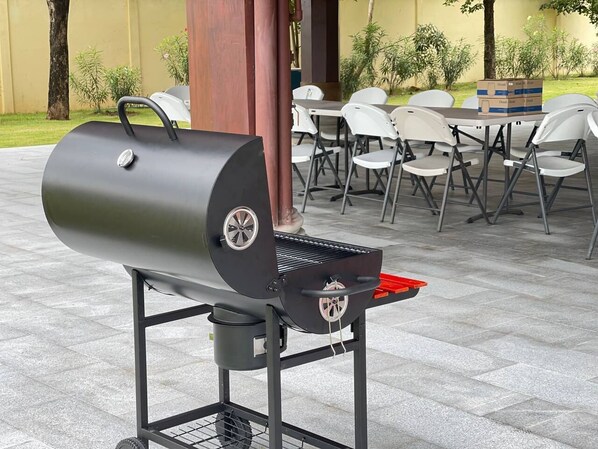 A portable grill with wheels, a metal stand, and a red tray, set up outdoors with white chairs and tables in the background.