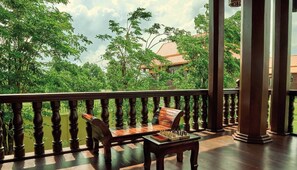 Une terrasse en bois avec un jeu d’échecs, un banc et une vue sur une végétation luxuriante et un bâtiment traditionnel.