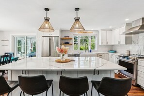 Une cuisine moderne dotée d’un grand îlot central, de meubles blancs, d’appareils électroménagers en acier inoxydable et de luminaires suspendus.