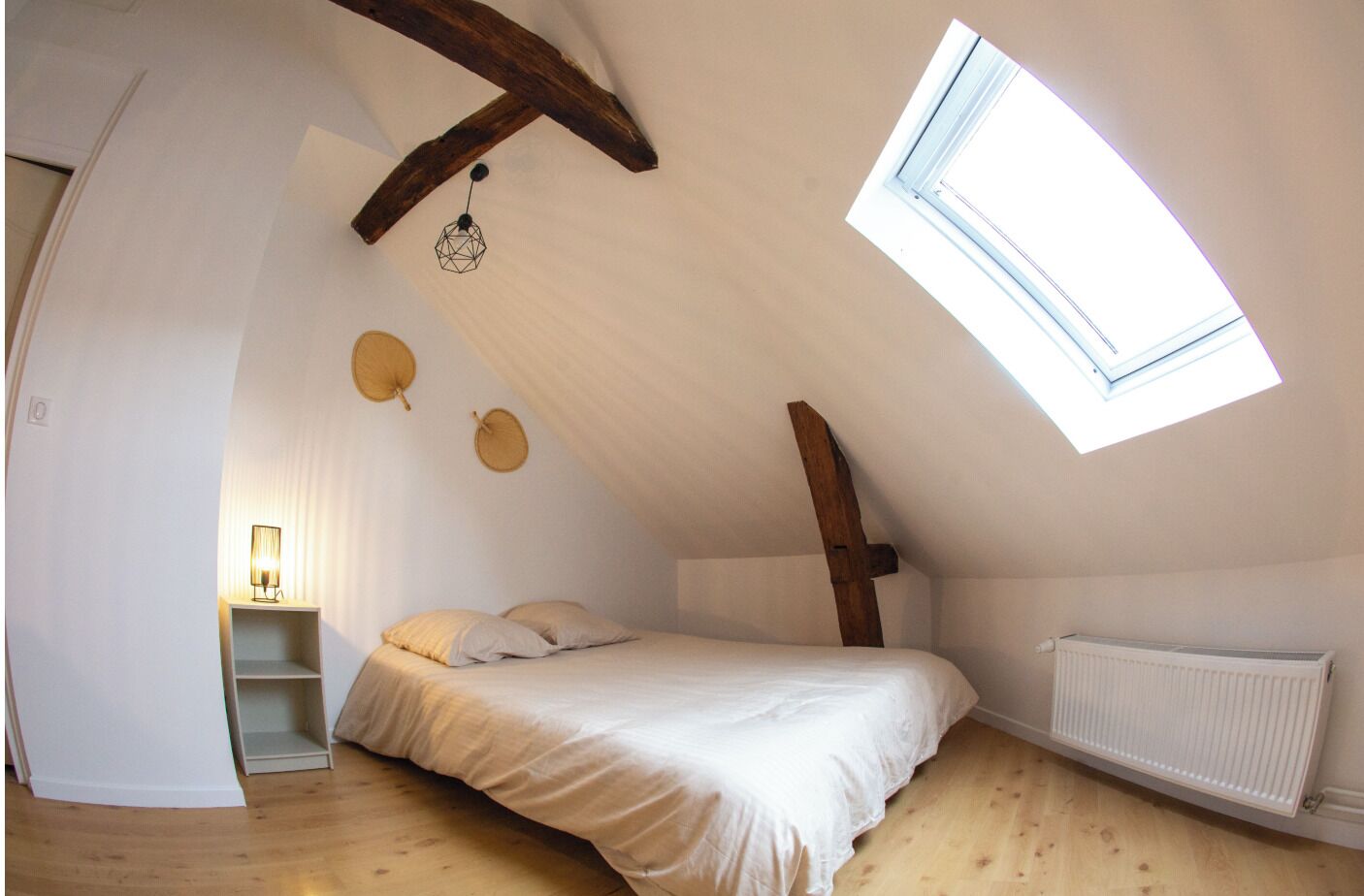 Un dormitorio con techo inclinado, tragaluces, cama con almohadas, mesita de noche con lámpara y radiador.
