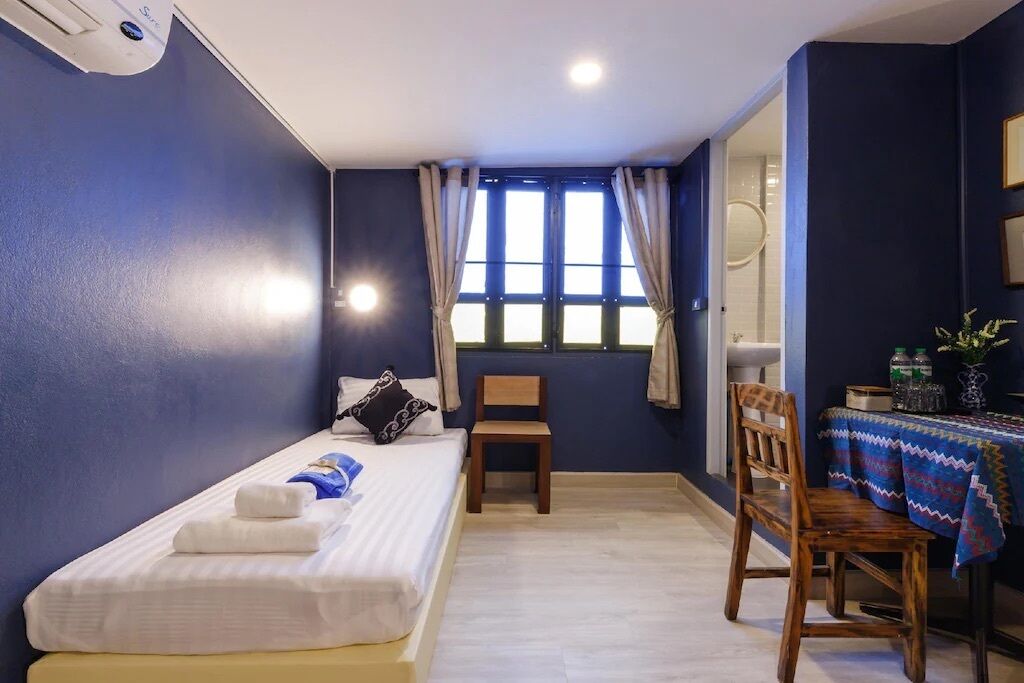 A bedroom with a single bed, a chair, a small table with a tablecloth, and a window with curtains.