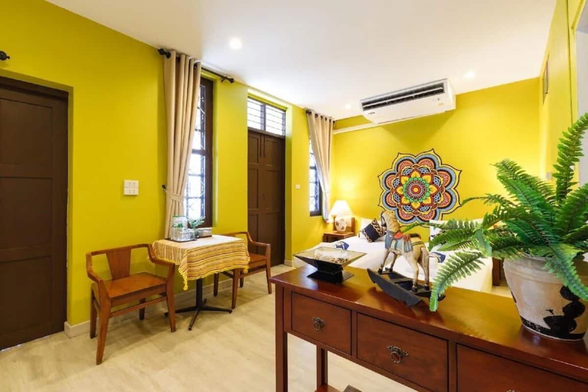 A room with yellow walls, a wooden desk, a chair, a bed with a colorful headboard, and a potted plant.