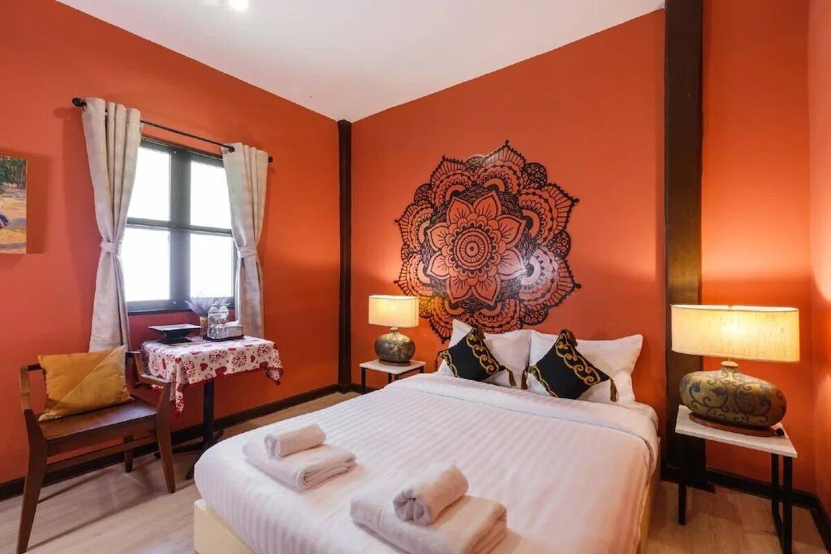 A hotel room with a bed, two bedside tables with lamps, a window with curtains, a small table with a lamp, and a chair with a cushion.