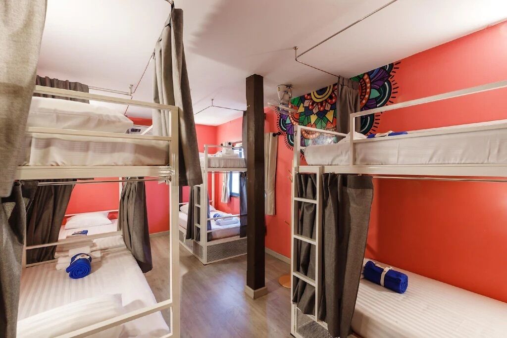 A dormitory room with bunk beds, a colorful tapestry on the wall, and a small window.