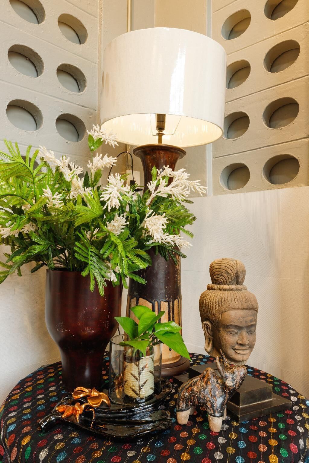 A lamp with a white shade, a potted plant with green leaves and white flowers, and a small elephant figurine on a round table with a colorful patterned tablecloth.