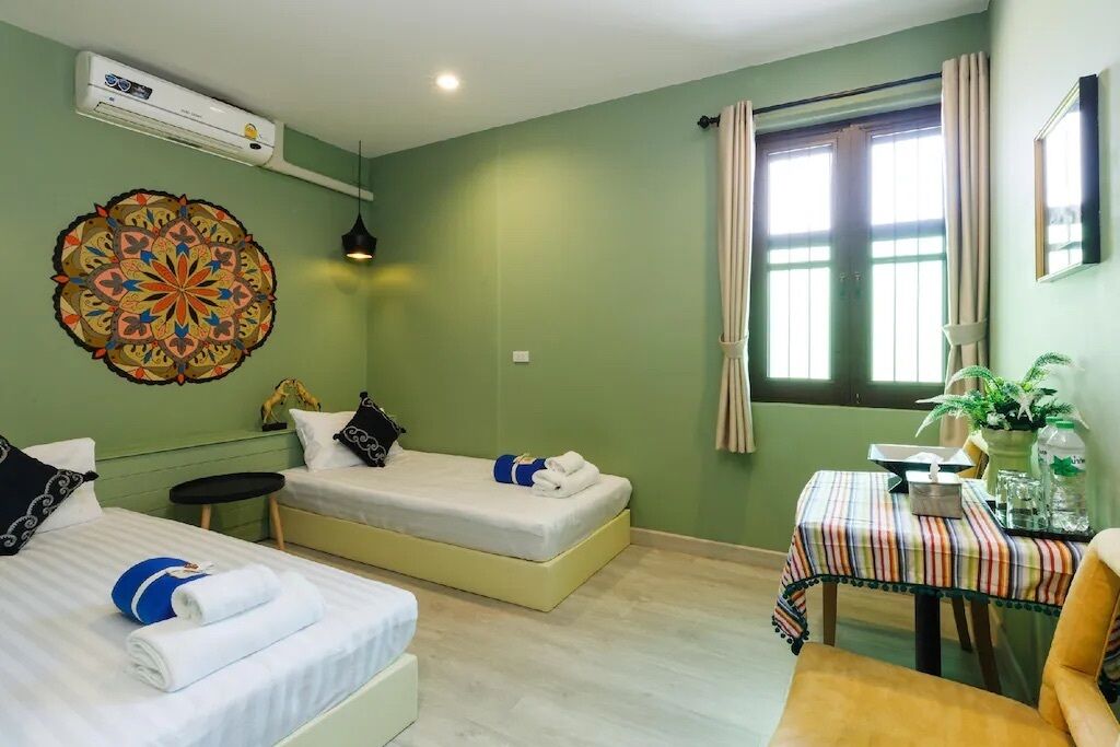 Una habitación con dos camas, una mesita con mantel a rayas, una silla, un adorno de pared y una ventana con cortinas.