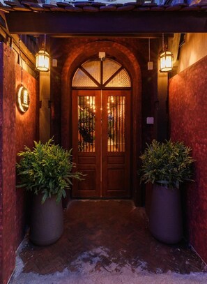 Uma entrada em arco de madeira com porta de vidro, ladeada por dois grandes vasos de plantas e iluminada por lanternas penduradas.