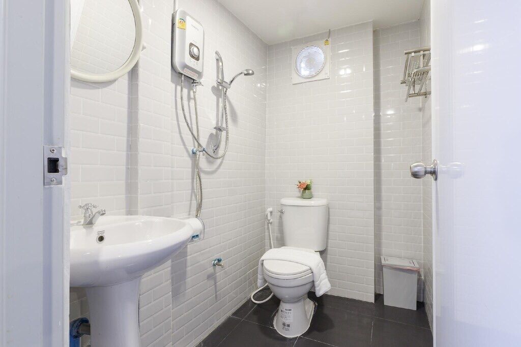 A small, clean bathroom with white tiles, a white sink, a toilet, and a shower unit.